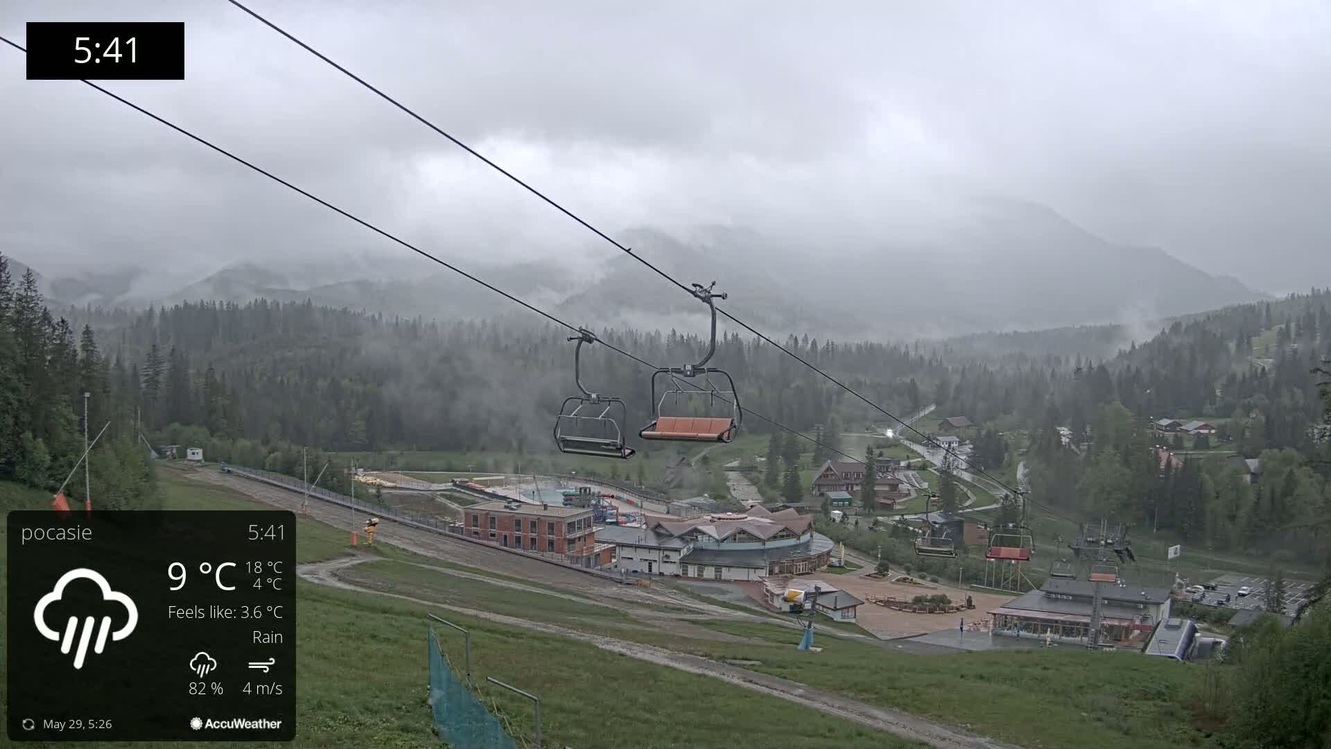 Two empty chairlifts hang against a rainy, overcast sky above a mountain village nestled among misty, dark green forests.