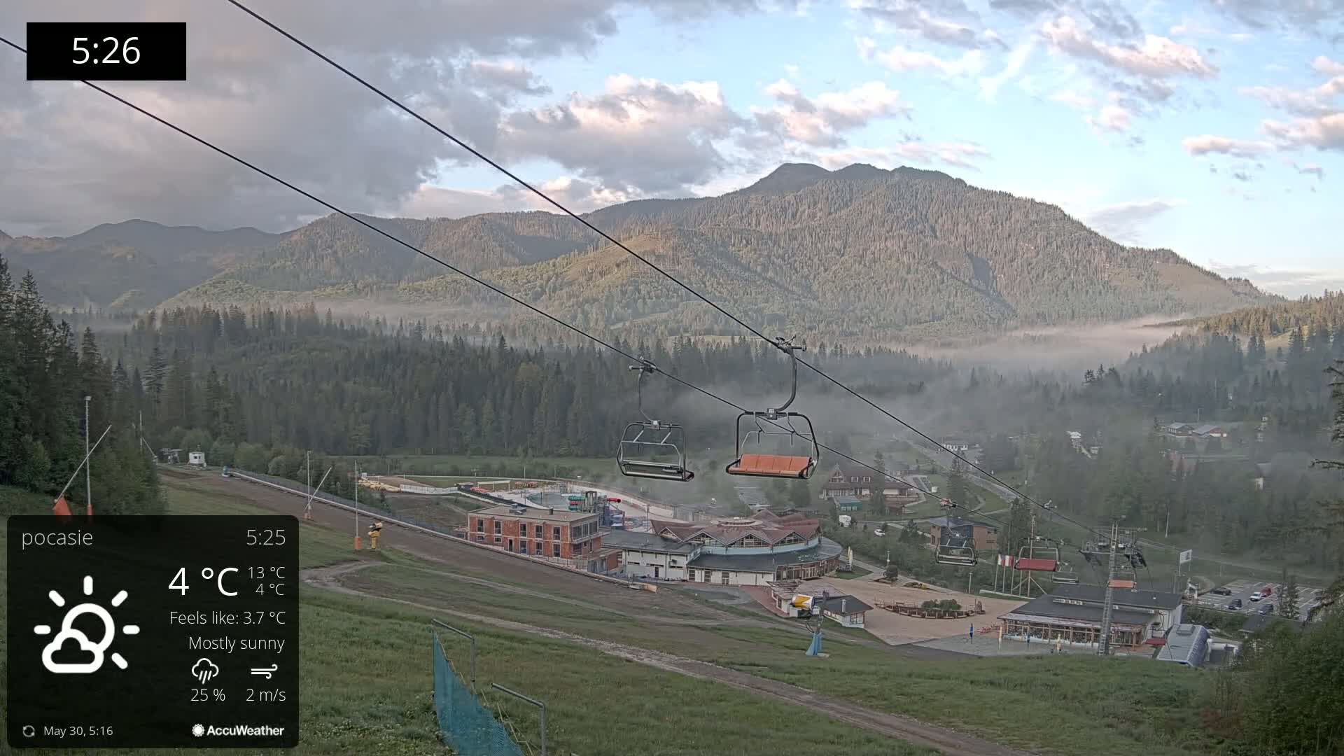 A mostly sunny mountain valley scene with low-lying fog, featuring a chairlift and resort buildings at the base of the mountains.