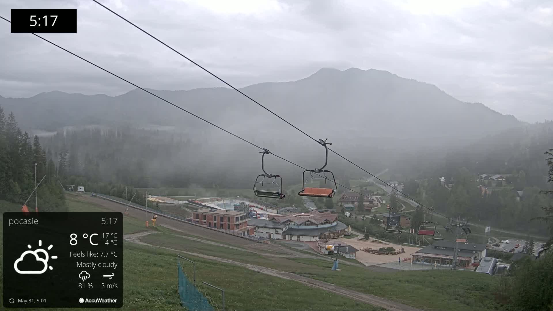 Two empty chairlifts hang from cables above a mostly cloudy mountain valley scene with low-hanging fog.