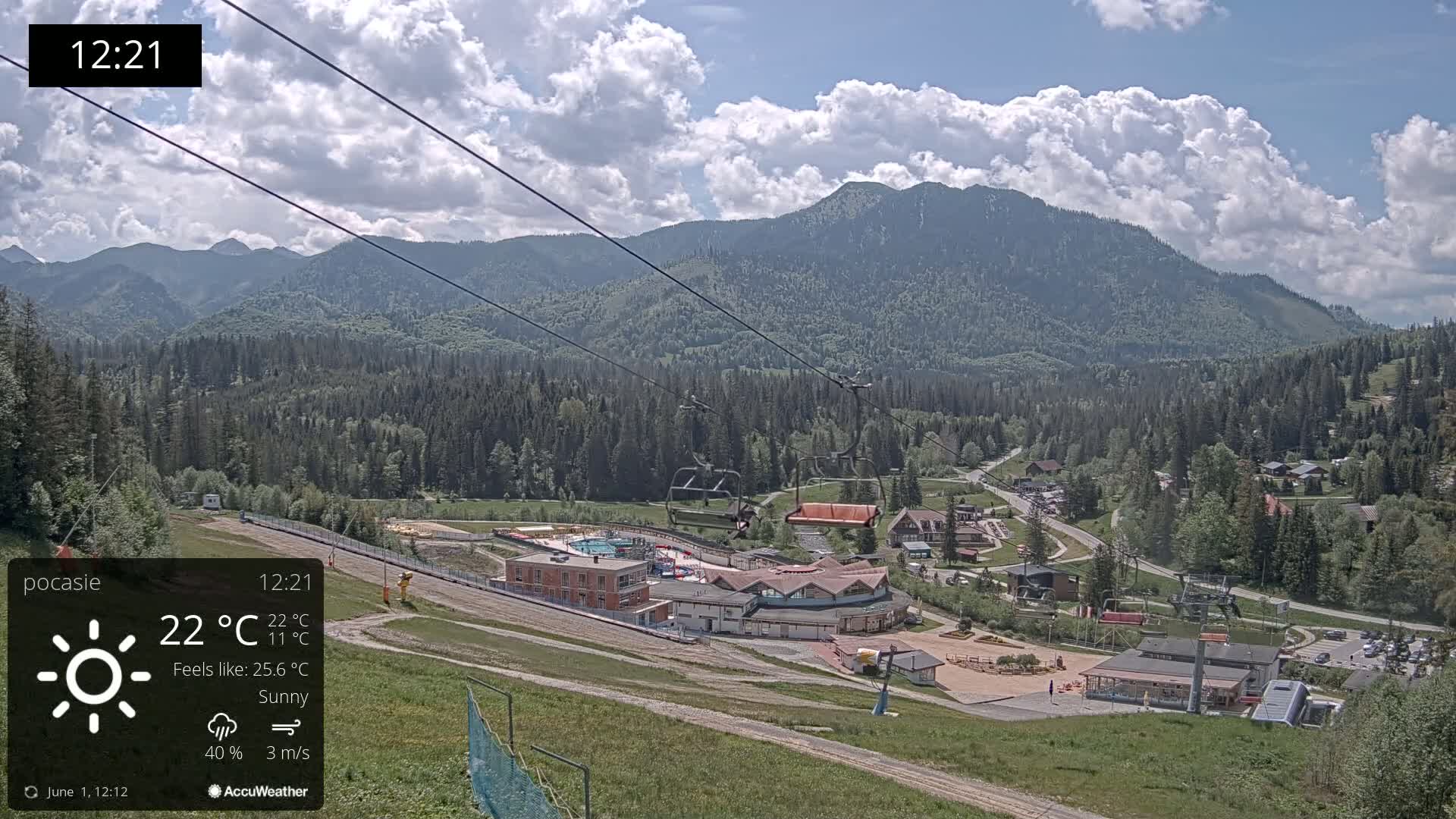 A sunny mountain resort with a chairlift and buildings nestled in a valley, under a partly cloudy sky.