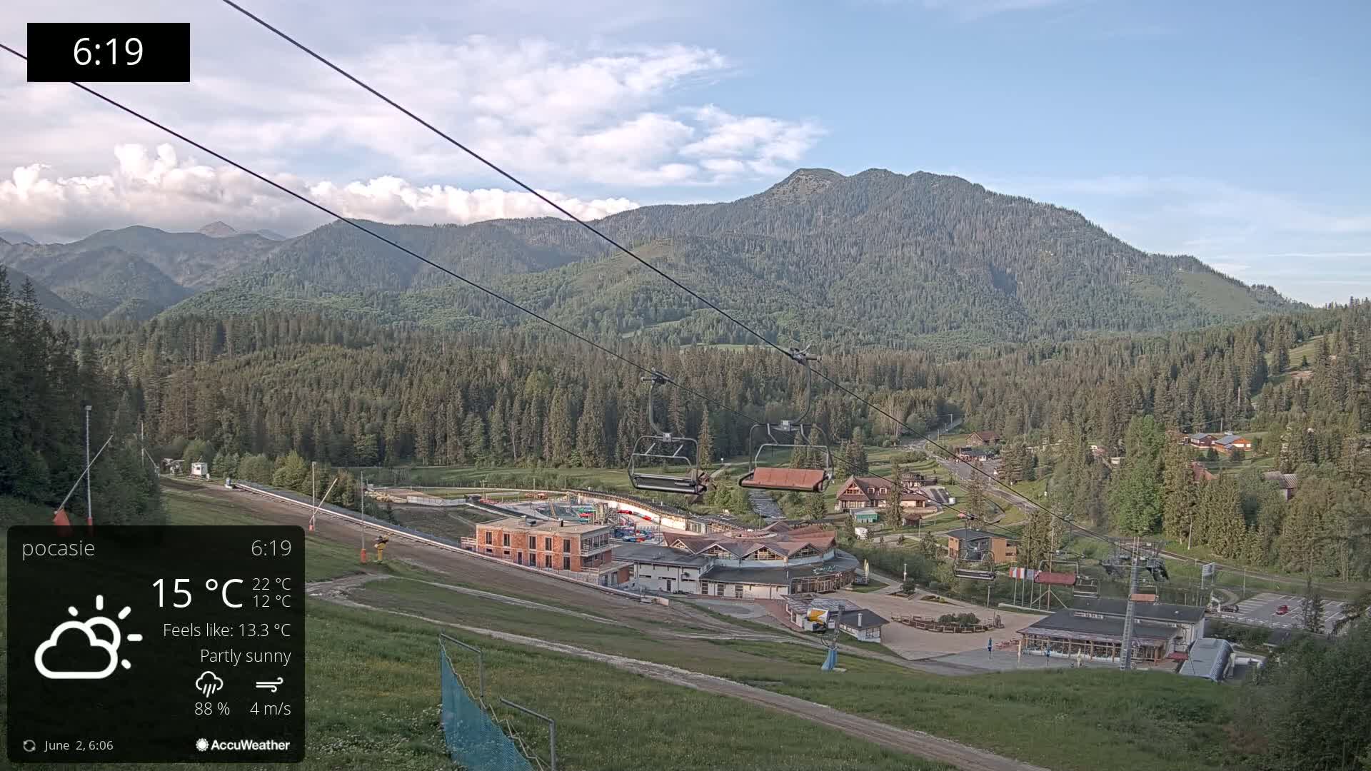 A partly sunny mountain scene shows a ski lift ascending a grassy hill toward a complex of buildings nestled in a forest at the base of a mountain range.
