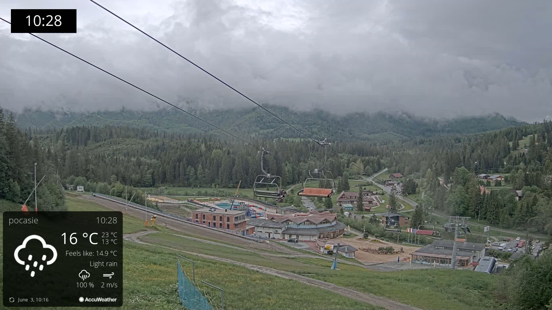 A chairlift ascends a mountainside overlooking a village nestled in a valley under a cloudy sky with light rain.
