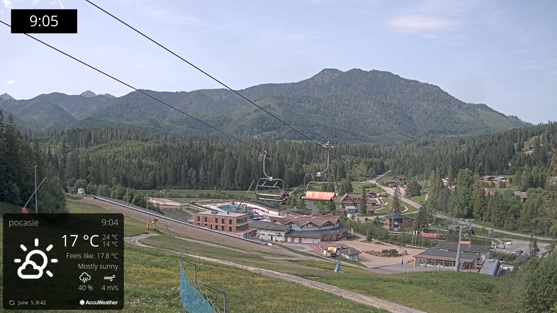 A mostly sunny mountain view shows a ski lift ascending toward a village nestled in a forest at the base of a mountain range.