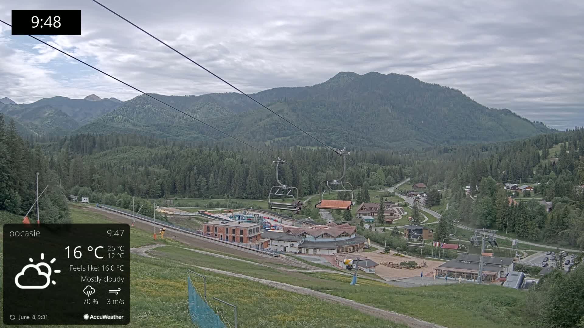 A mostly cloudy mountain landscape is viewed from a ski lift, with a village and buildings below.