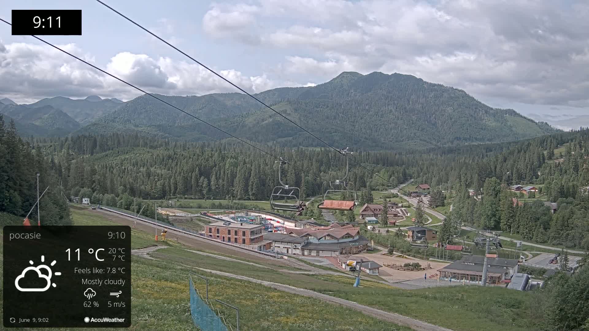 A mostly cloudy mountain vista with a ski resort below, featuring a chairlift and several buildings under a light breeze.