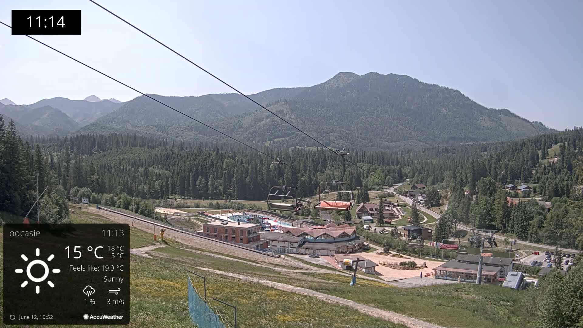 A sunny mountain vista shows a ski lift ascending a hillside toward a resort complex nestled among evergreen trees and mountains.