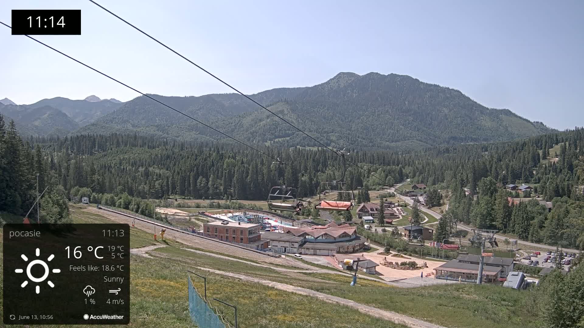 A sunny mountain resort village with a chairlift and swimming pool, nestled amongst green forested hills under a clear sky.