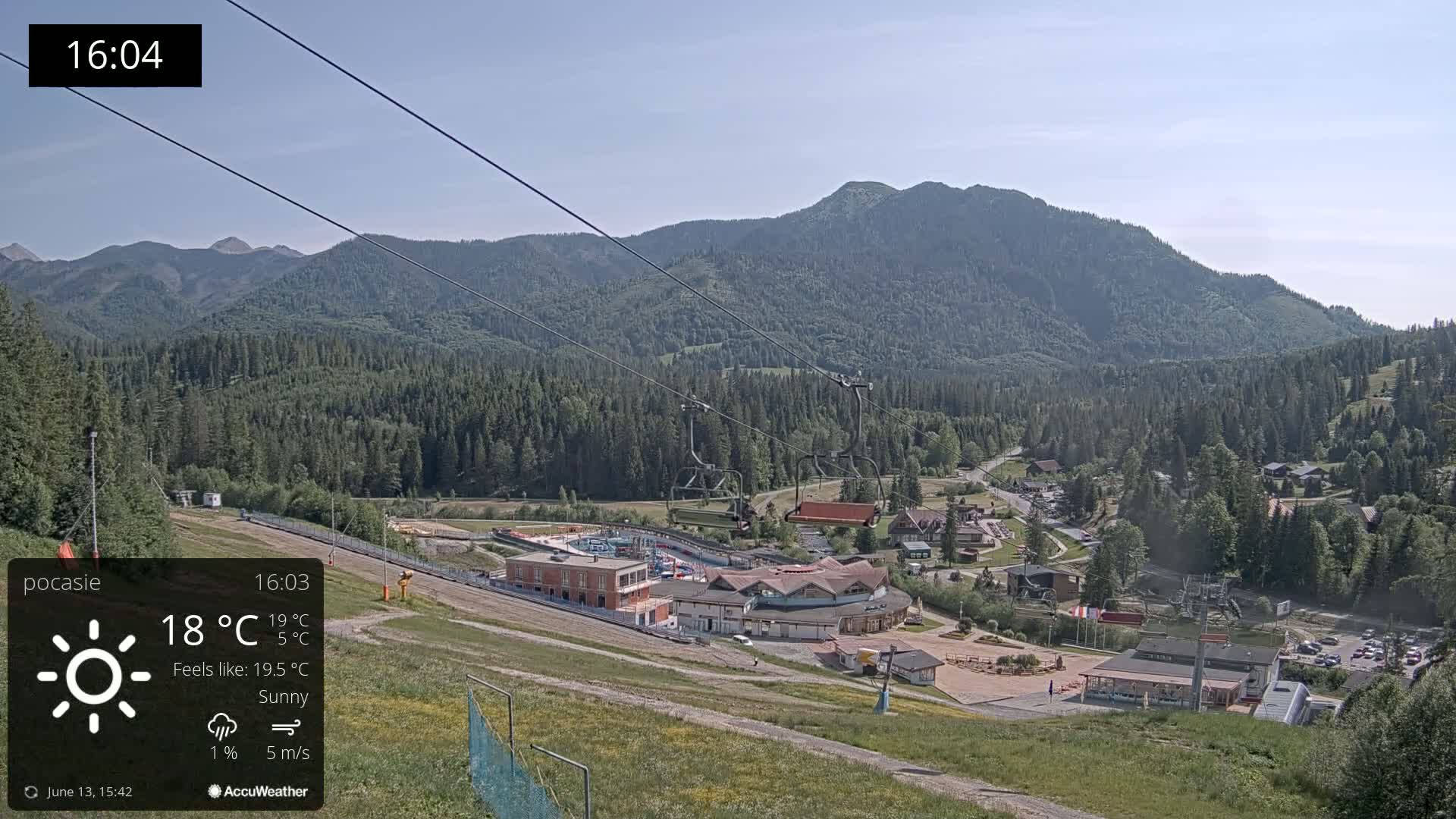 A sunny mountain resort with a chairlift and several buildings is visible on a hillside, under a clear sky.