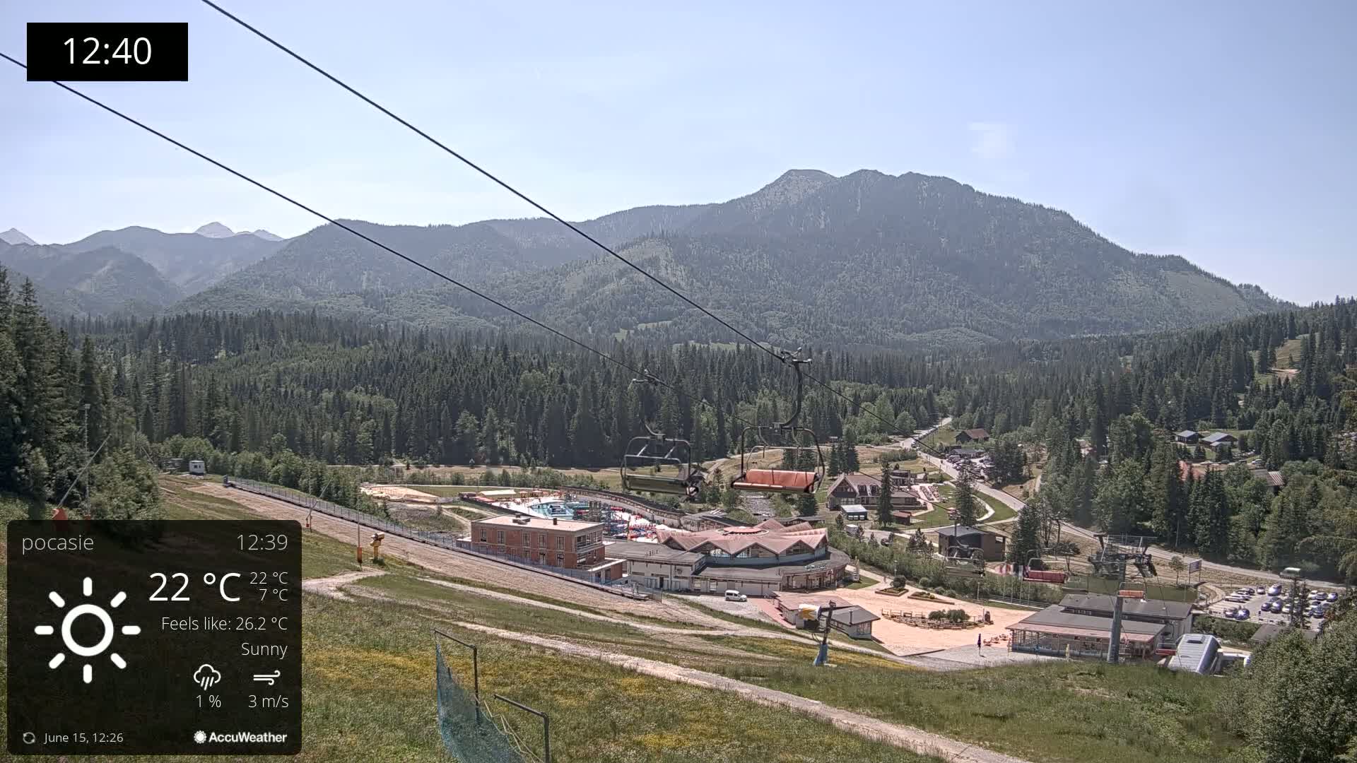 A sunny mountain resort scene with a chairlift and buildings below, viewed from a hillside on a clear day with a light breeze.