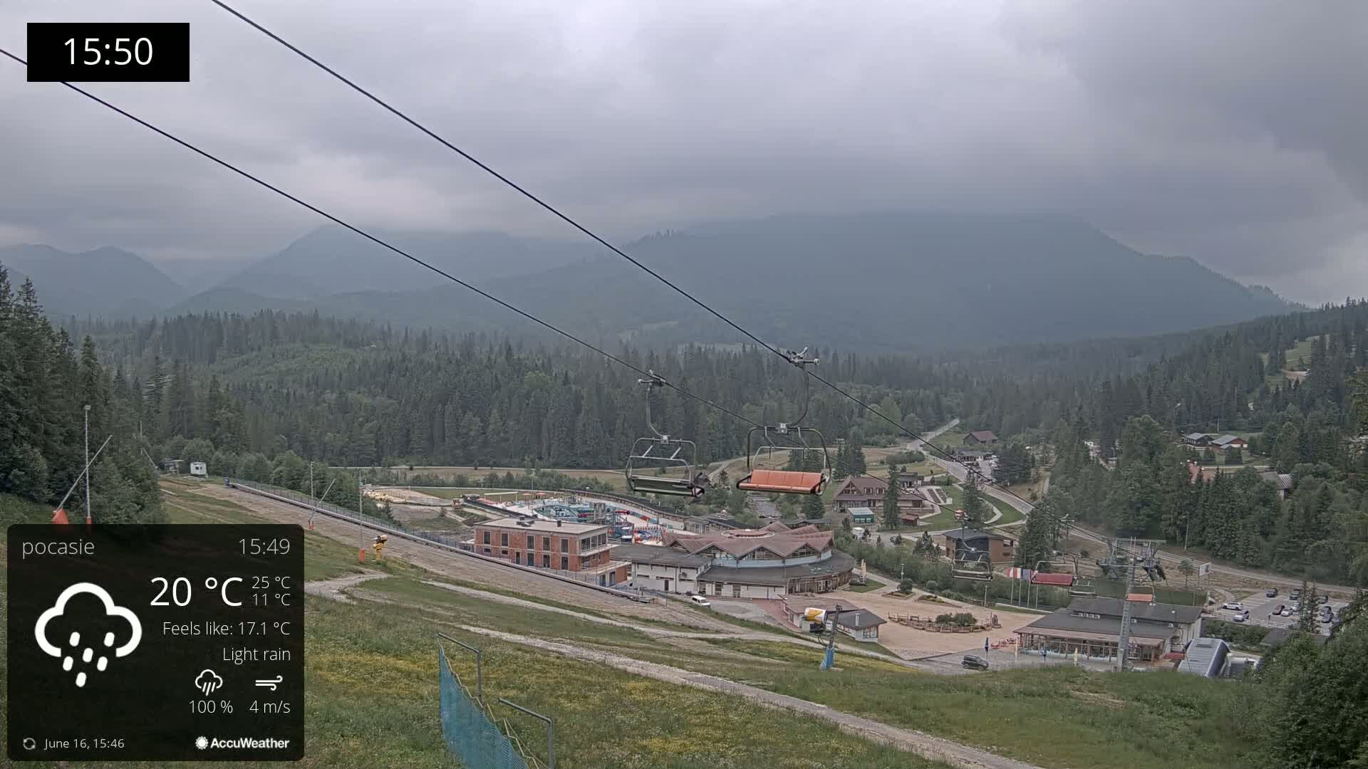A chairlift ascends a mountainside toward a village nestled in a valley under a cloudy sky with light rain.