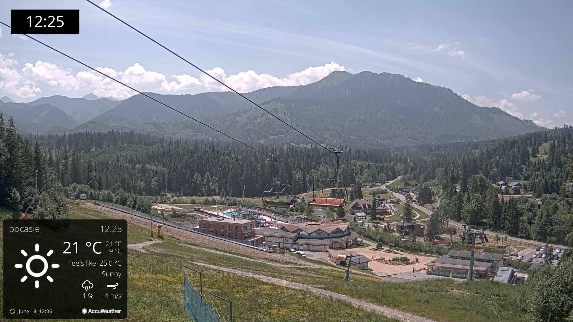 A sunny mountain resort scene with a chairlift and buildings, under a mostly clear sky with a few clouds.