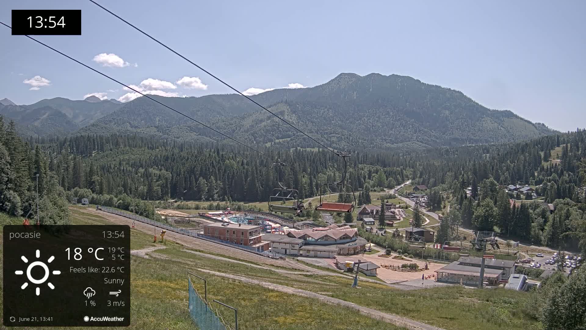 A sunny mountain resort with a chairlift, buildings, and a swimming pool is nestled amongst green hills and a large mountain range under a clear blue sky.