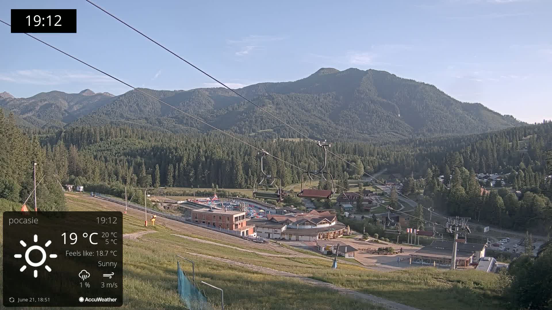 A sunny mountain resort with chairlifts and buildings is nestled among lush green forests under a clear blue sky.