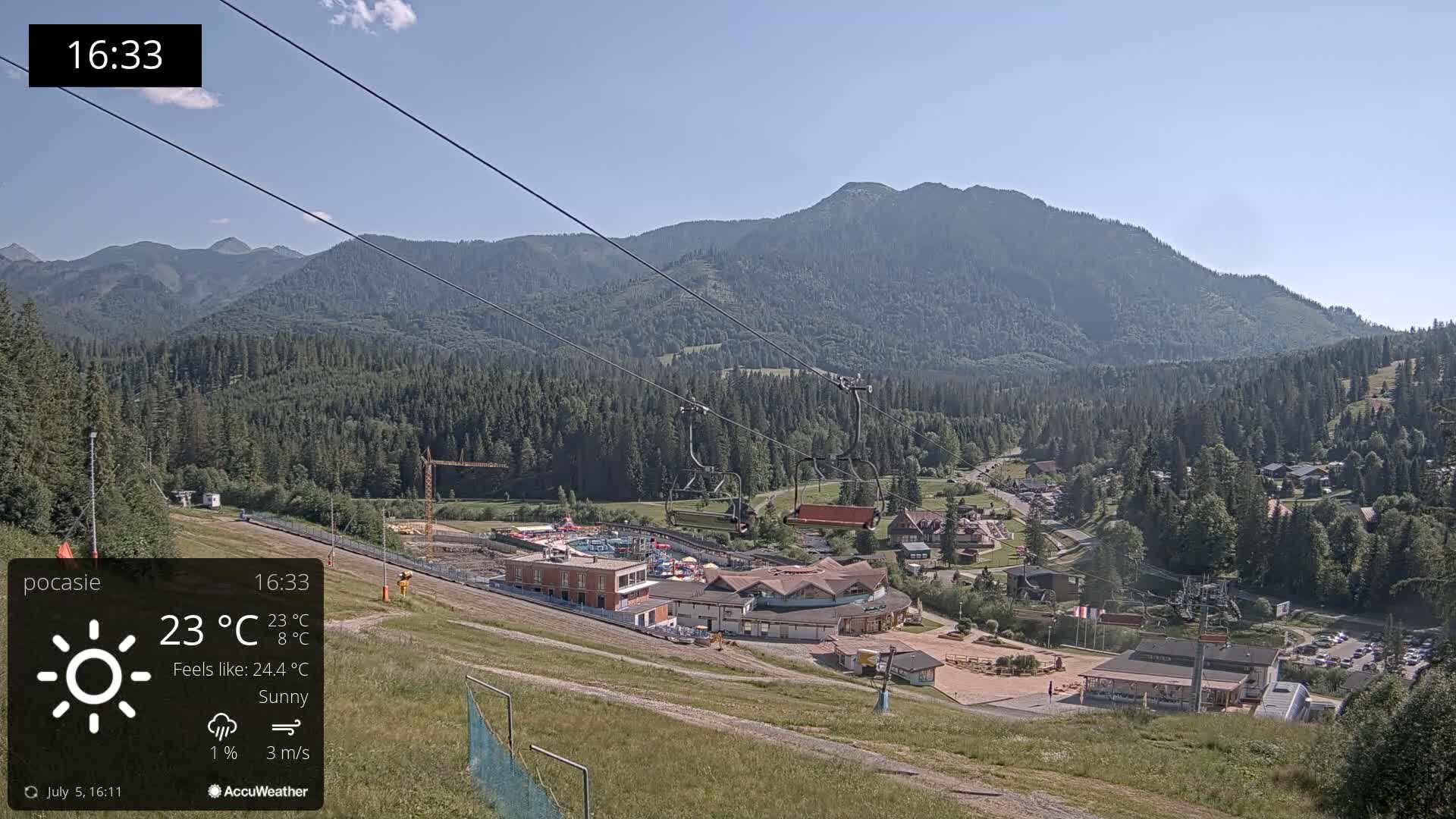 A sunny mountain resort scene with a chairlift, buildings, and a large mountain in the background.