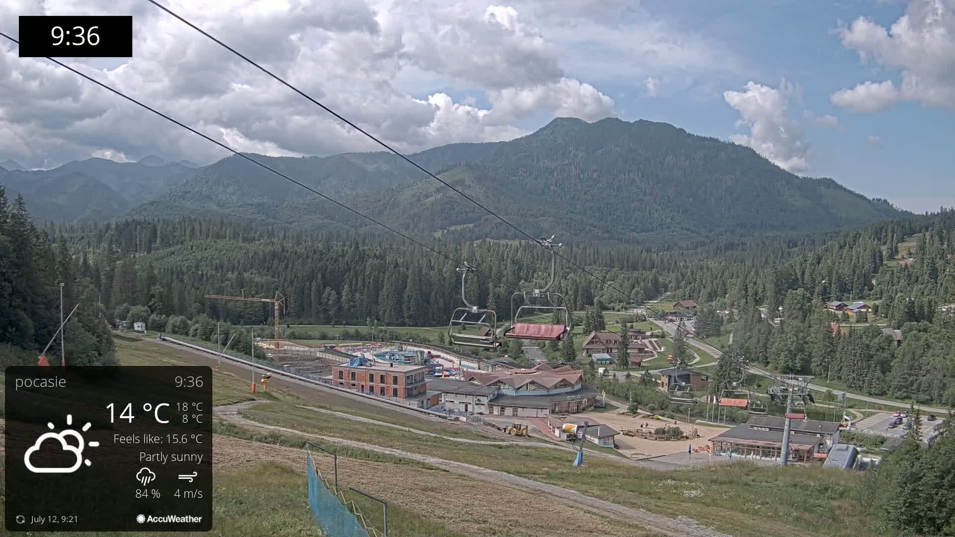 A partly sunny mountain vista shows a chairlift ascending towards a ski resort nestled amongst a forest, with a small village visible in the valley below.