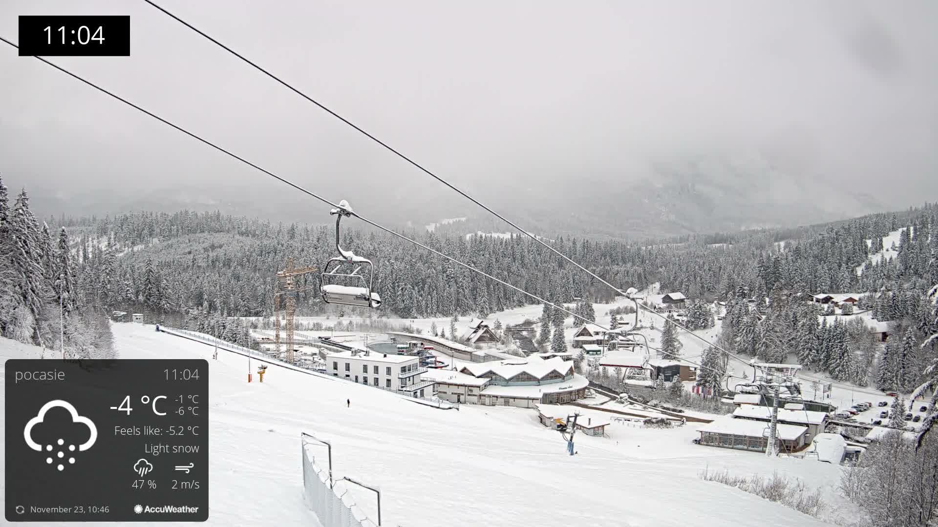 A snowy mountain ski resort is seen under an overcast sky with light snow falling, featuring ski lifts traversing over snow-covered slopes, dense pine forests, and scattered buildings nestled in the valley.