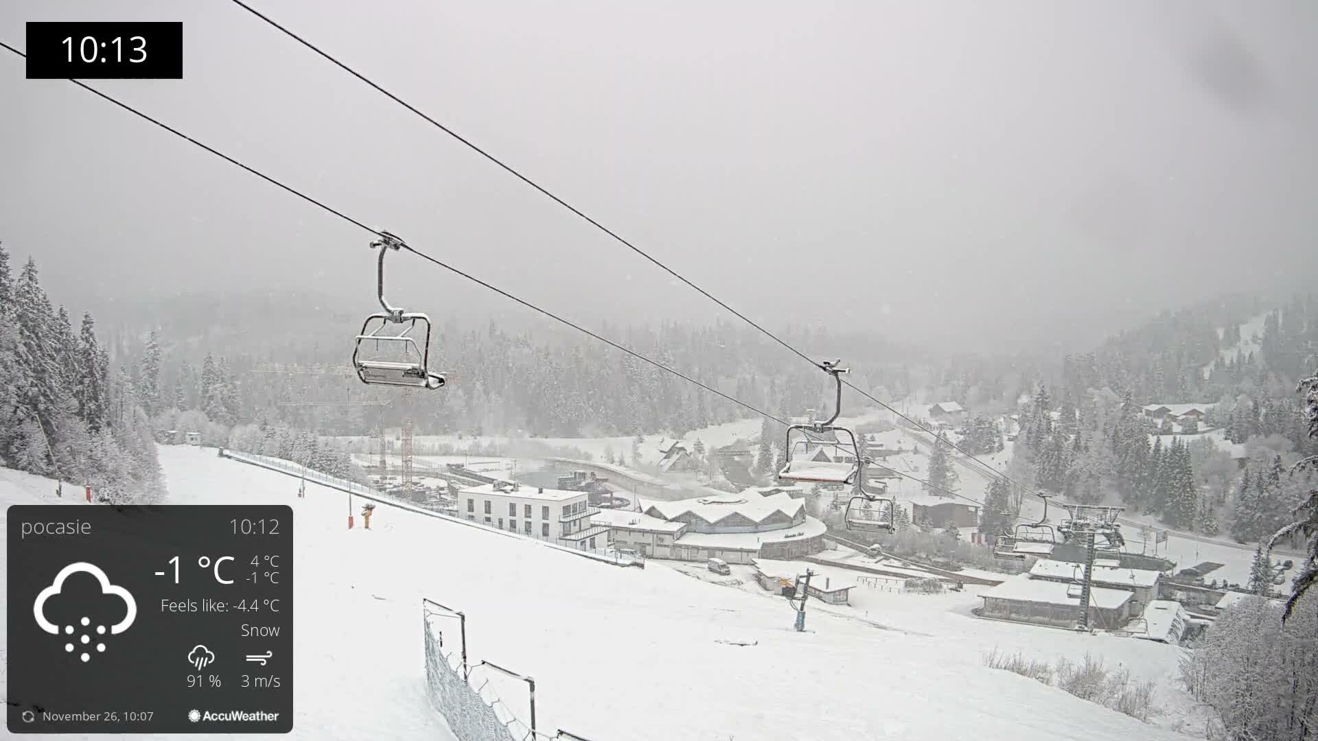 A snow-covered mountain ski resort is seen under heavy snowfall and overcast skies, featuring empty chairlifts traversing over pine trees and buildings blanketed in white.