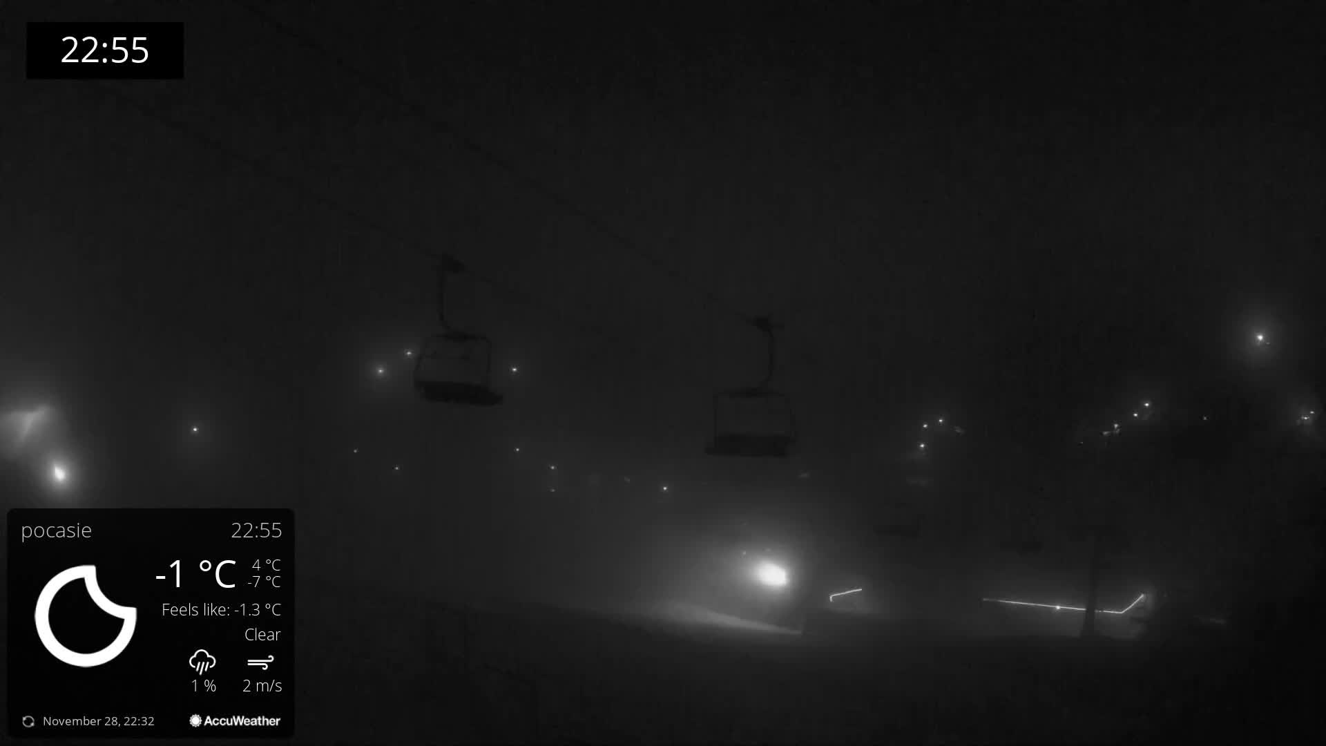 The image depicts a dark and very foggy winter night at a ski resort, with two chairlifts hanging above a snow-covered ground illuminated by several distant lights and a prominent bright light in the lower right.