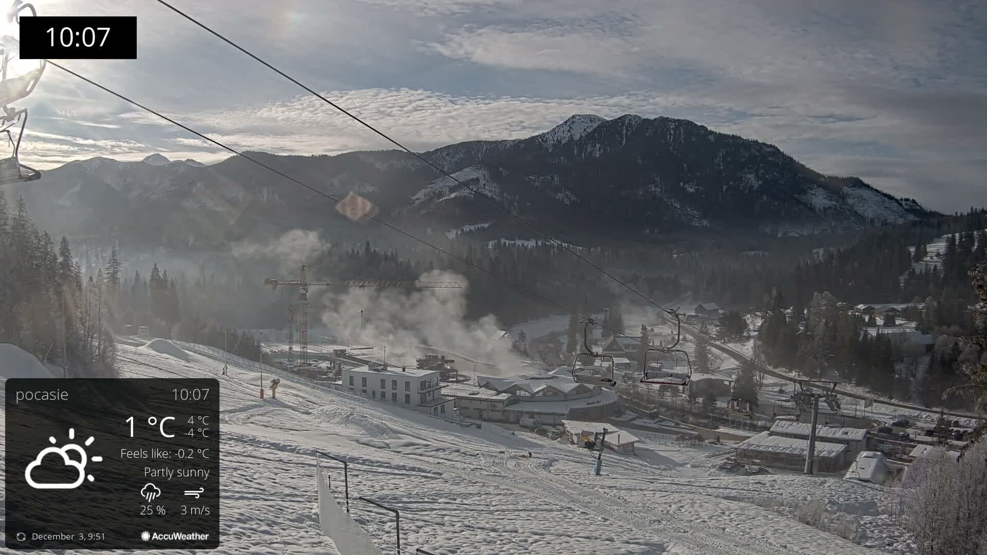 A snow-covered mountain ski resort is pictured under a partly sunny sky, showing active snow-making operations near buildings and chairlifts, with dense evergreen forests and hazy, snow-capped peaks in the background.