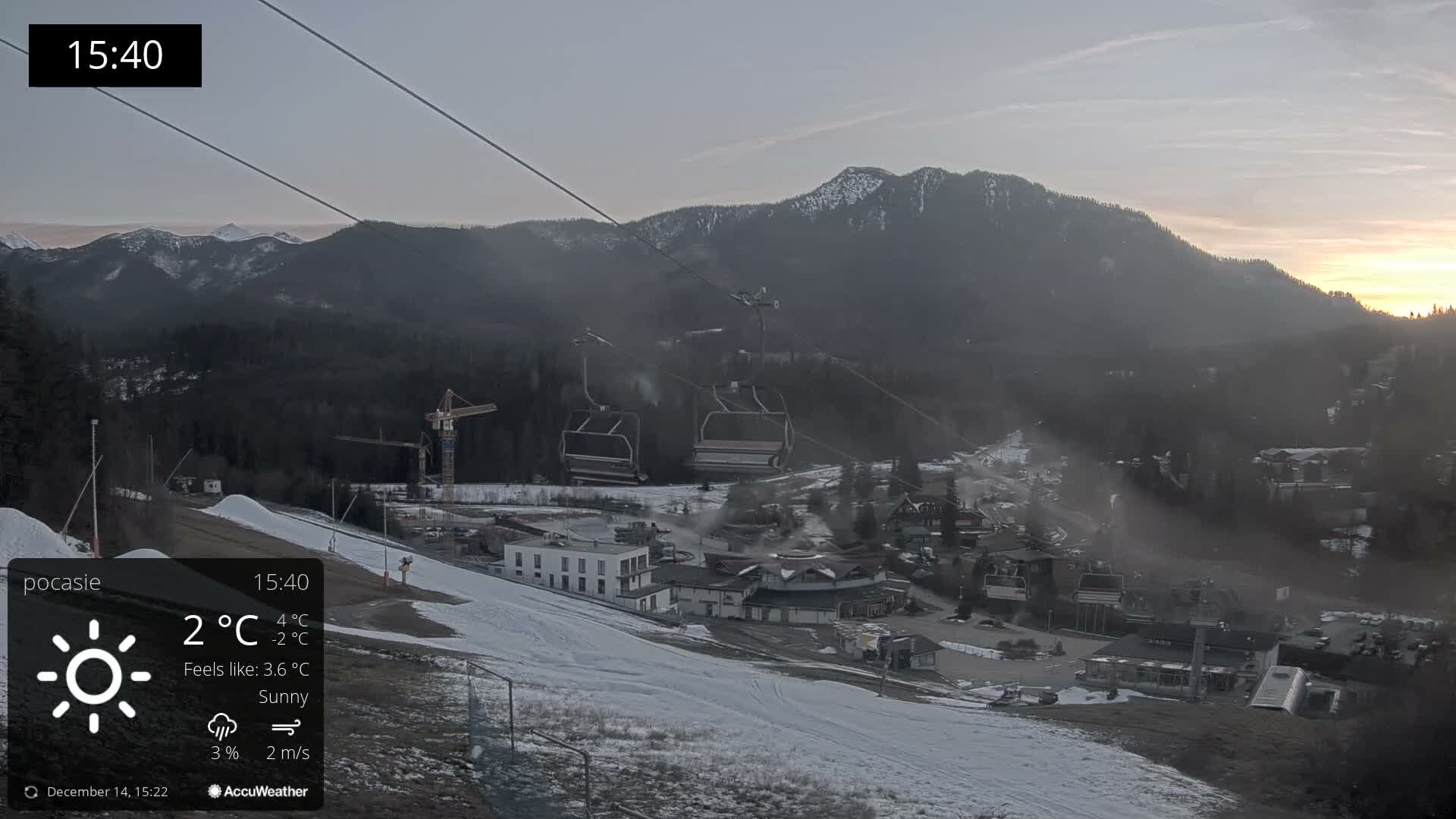 A snow-covered mountain ski resort is pictured under a partly sunny sky, showing active snow-making operations near buildings and chairlifts, with dense evergreen forests and hazy, snow-capped peaks in the background.