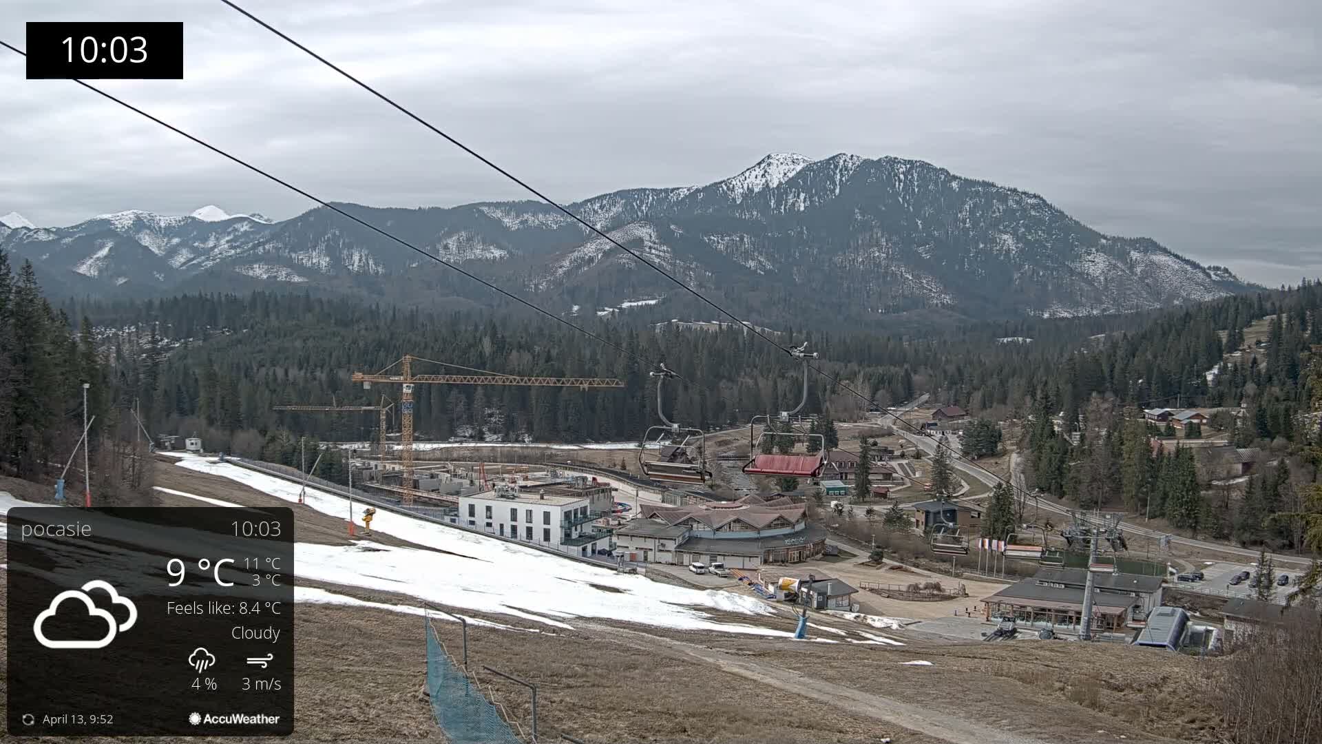 A snow-covered mountain ski resort is pictured under a partly sunny sky, showing active snow-making operations near buildings and chairlifts, with dense evergreen forests and hazy, snow-capped peaks in the background.