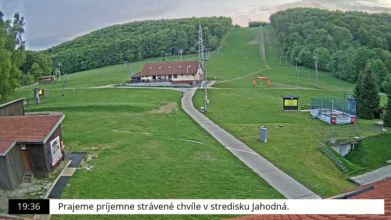 A ski resort with a building, grassy areas, ski lifts, and a paved pathway is visible under an overcast sky.