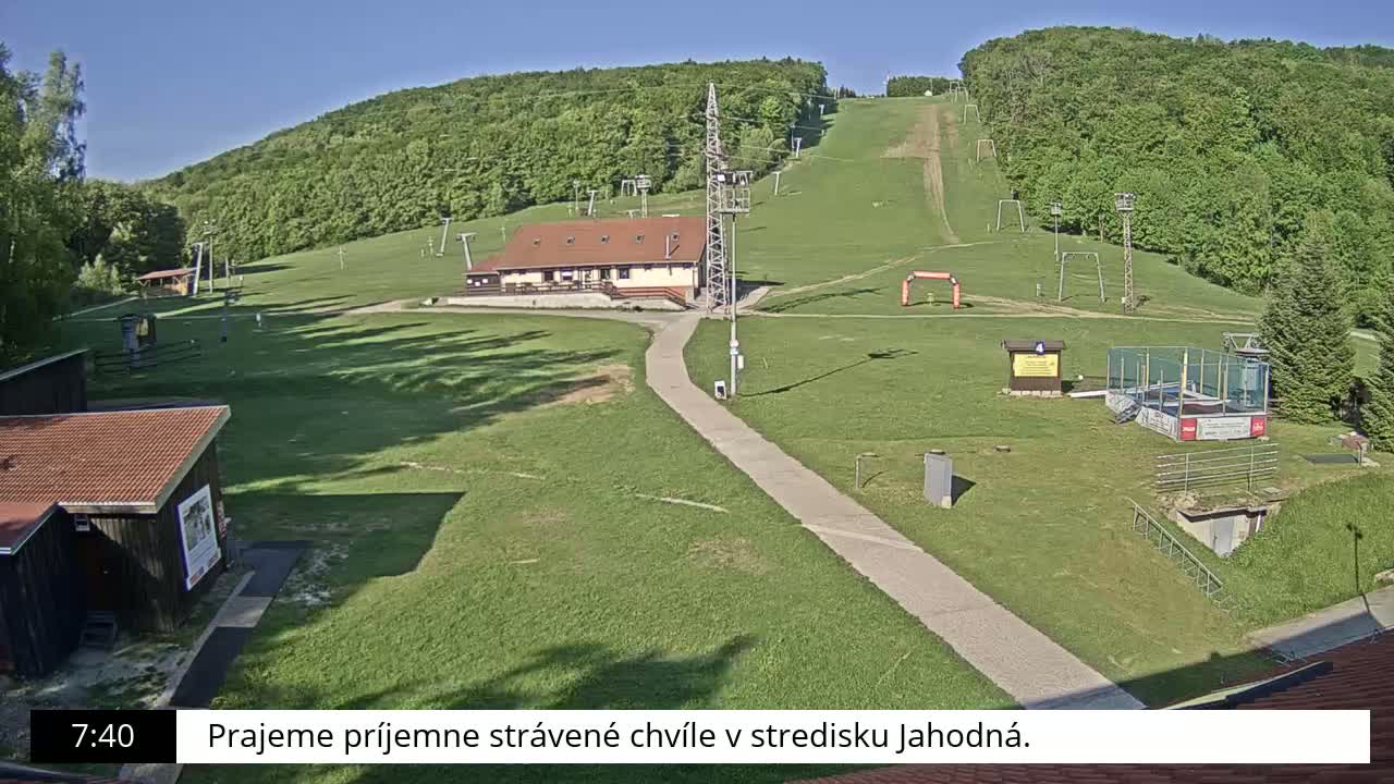 A ski slope with a building and several other structures is visible on a sunny day, surrounded by green grassy areas and a forest-covered hill.