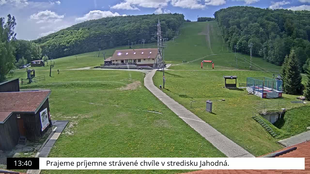 A grassy ski resort with a building, lift infrastructure, and a paved pathway is visible under a partly cloudy sky.