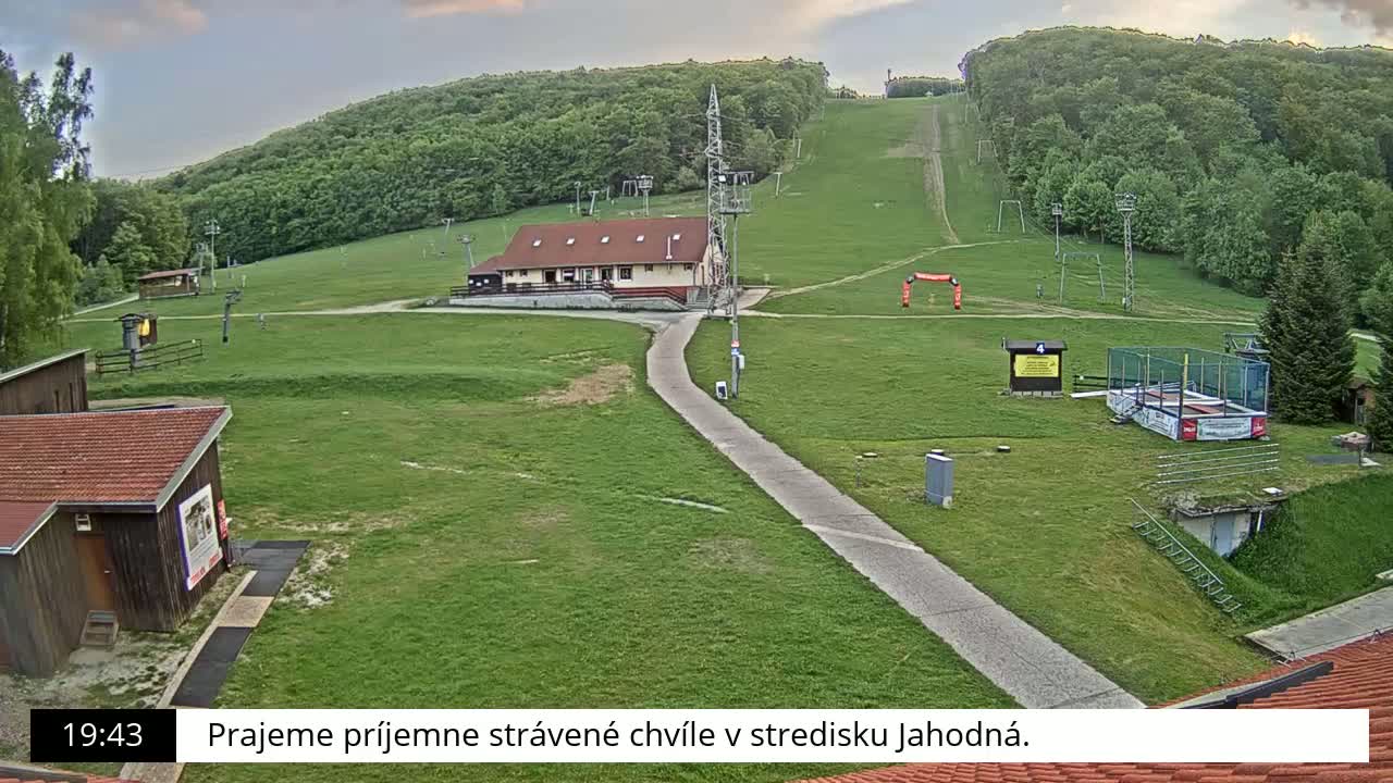 A grassy ski slope with a building, small structures, and a paved path is visible under a partly cloudy sky.