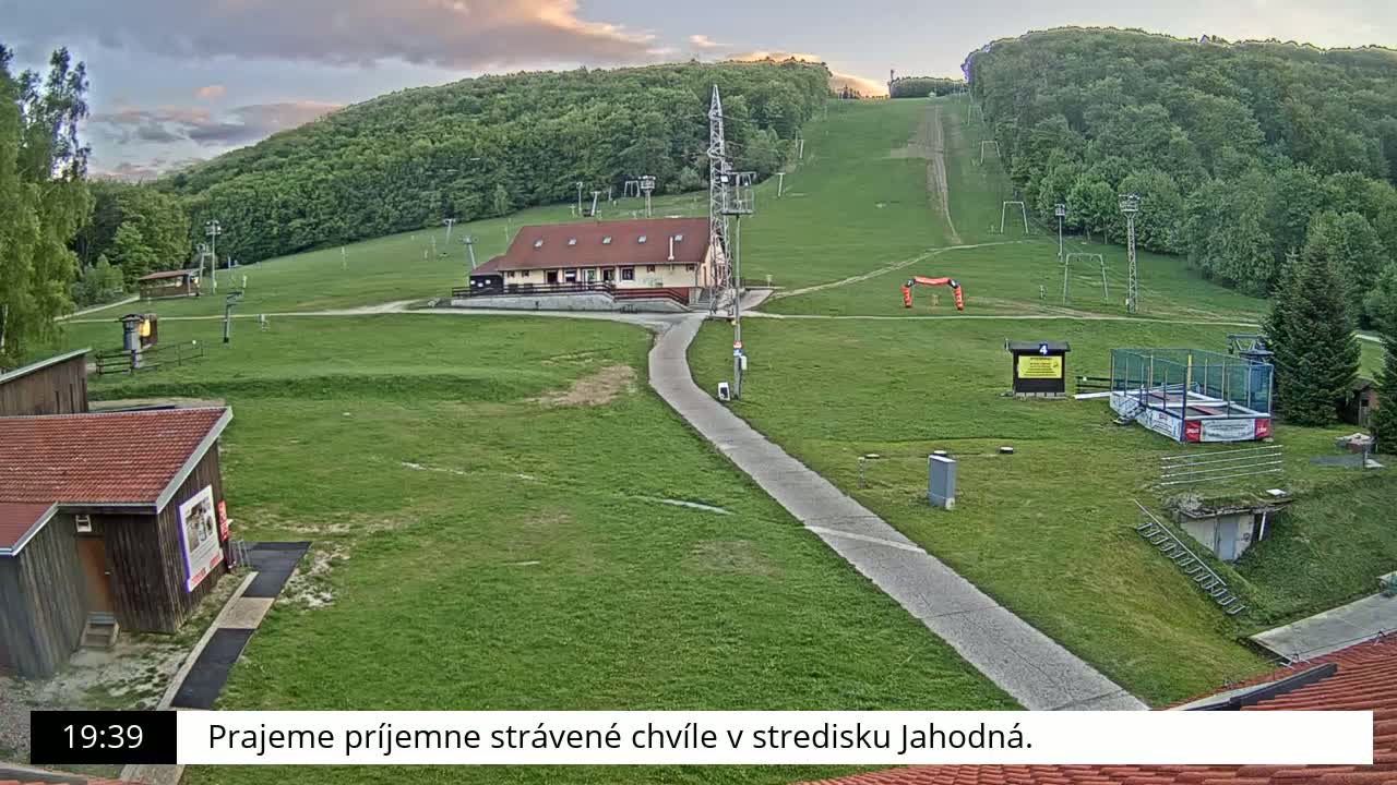 A grassy hillside with a ski lift, a building, and a paved pathway, under a partly cloudy sky.