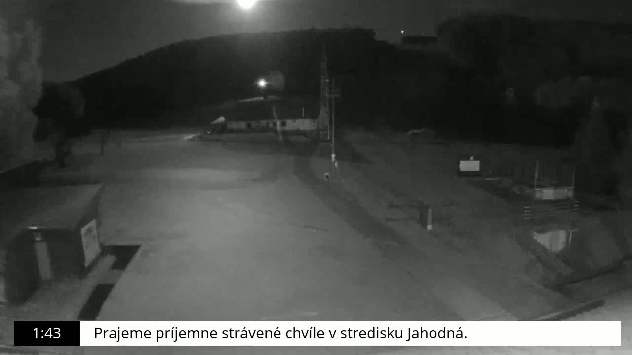 A nighttime, low-light black and white view of an empty paved area with a few structures and a hill in the background under a mostly clear sky.