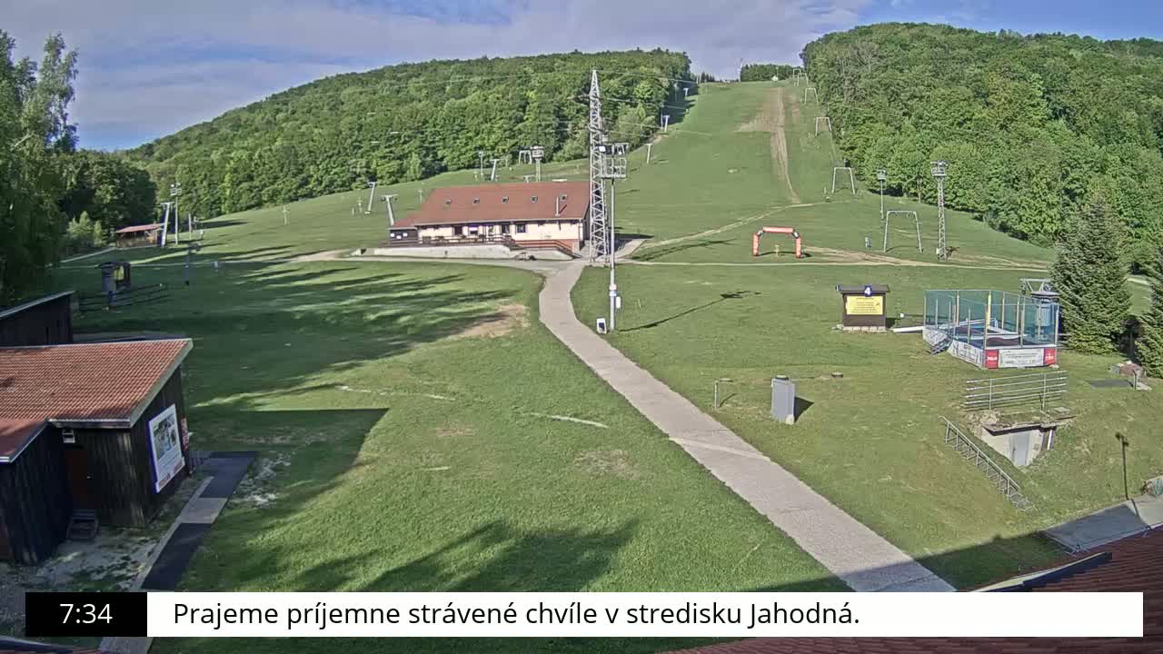 A ski slope with a building and lift equipment is visible on a sunny day, surrounded by green grass and trees.