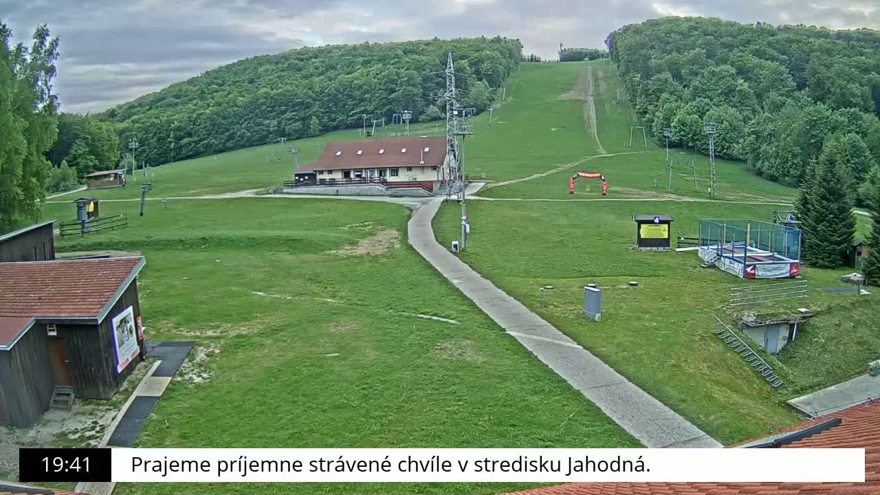 A grassy ski resort with a building, ski lifts, and a small trampoline is visible under a partly cloudy sky.