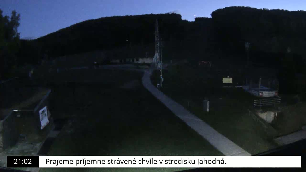 A paved path winds through a dark, grassy area at night, with a hill and a few buildings in the background under a twilight sky.