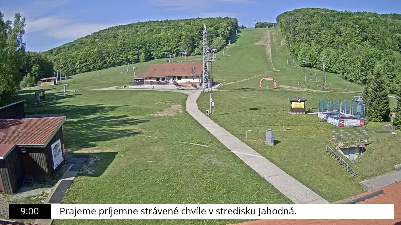 A paved path leads to a ski lodge nestled in a grassy area with ski lifts and a hillside covered in lush green trees under a sunny sky.
