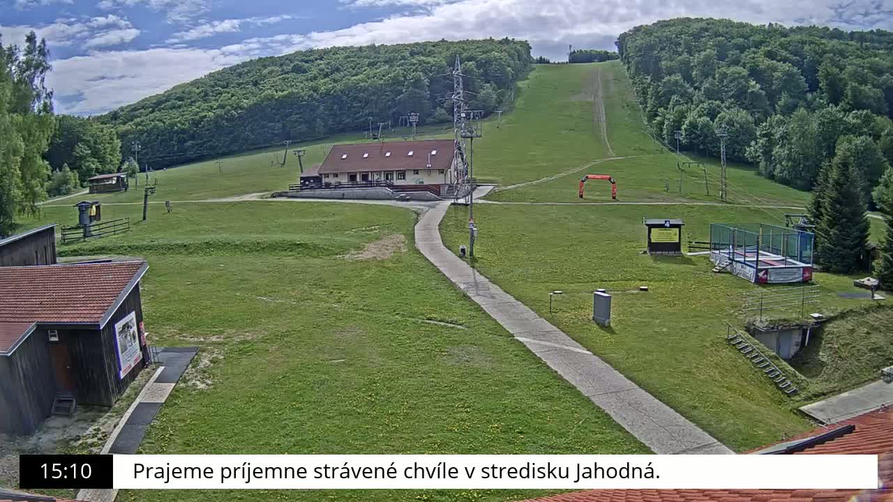 A grassy ski slope with a building, a paved pathway, and a trampoline is visible under a partly cloudy sky.