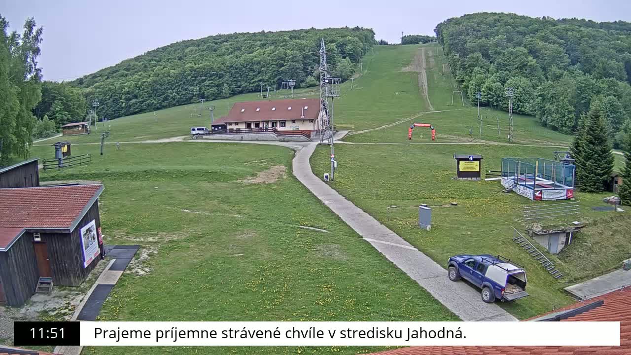 A ski slope with a building, some vehicles, and a trampoline sits on a grassy area below a hillside covered in green trees under an overcast sky.