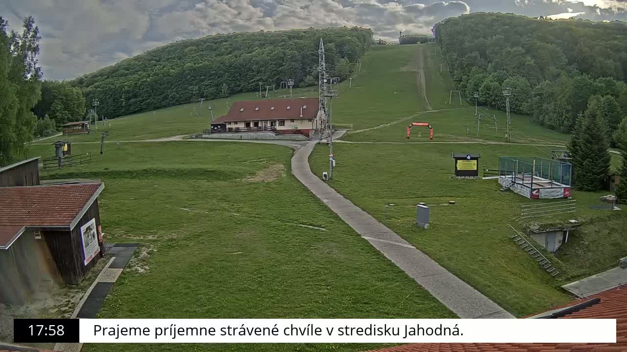 A grassy hill with a building, a paved pathway, and ski lifts visible under a partly cloudy sky.