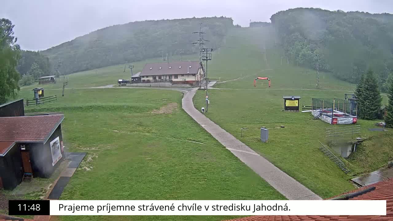 A grassy ski resort with a building and a paved pathway is seen on a rainy day, with hills in the background.