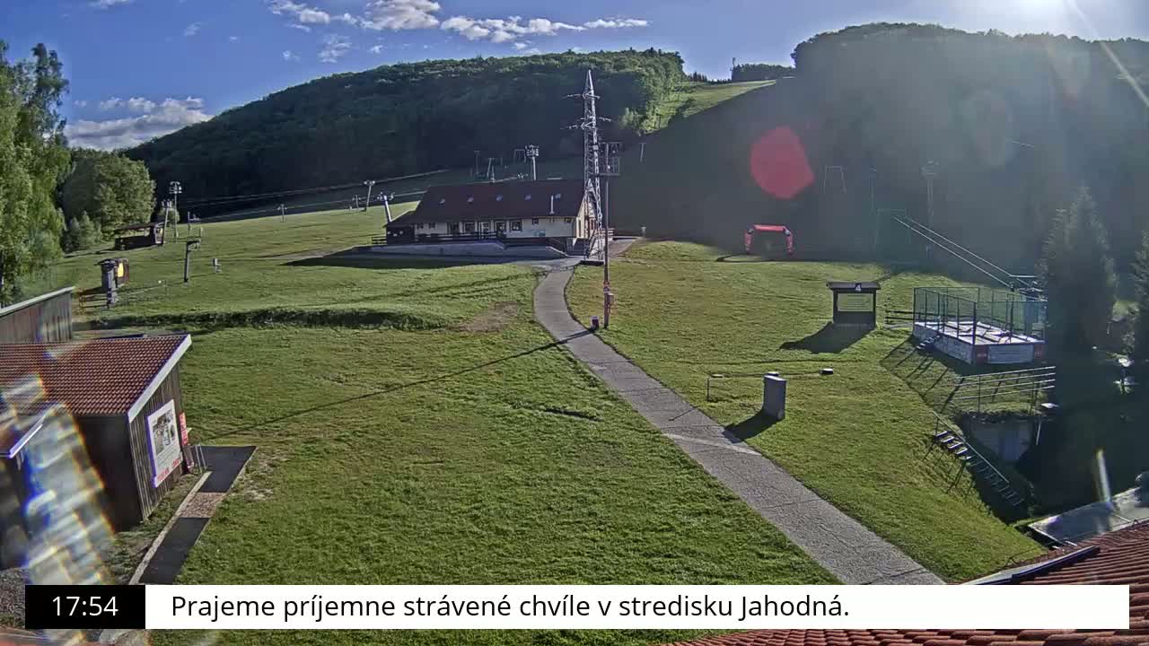 A sunny day at a ski resort shows a grassy area with buildings, a paved path, and ski lifts.