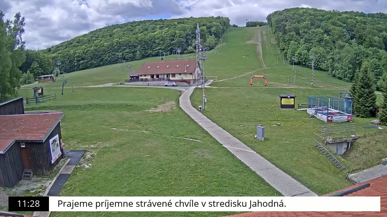A grassy ski slope with a building, a paved path, and some small structures is visible under a partly cloudy sky.