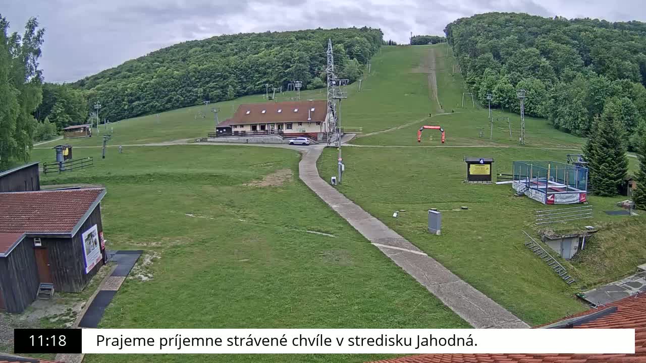 A grassy ski slope with a building, a paved path, and a trampoline under an overcast sky.