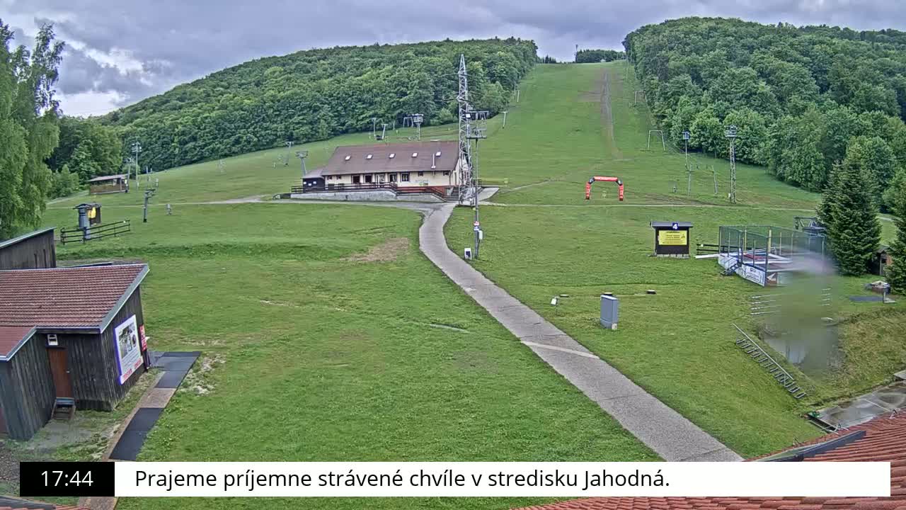 A grassy ski slope with a building and lift infrastructure is visible under a cloudy sky.