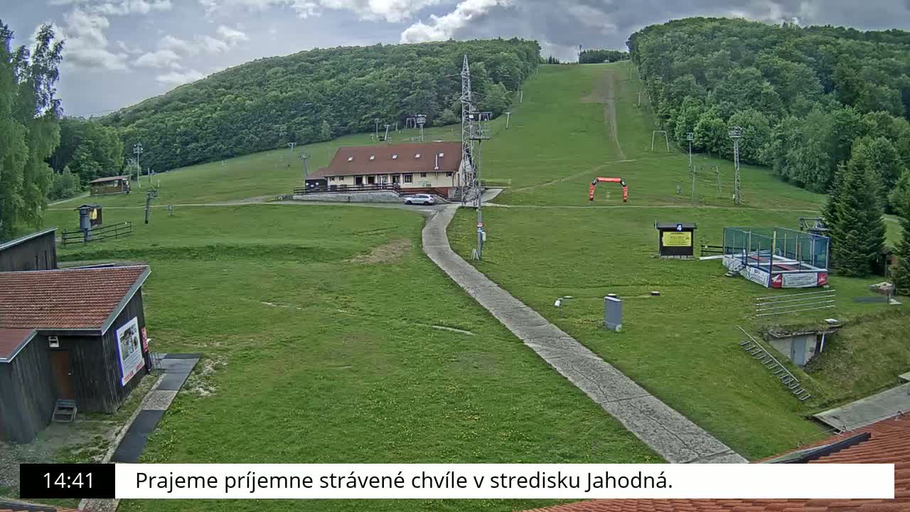 A grassy ski slope with a building, a paved path, and some recreational equipment is visible under a partly cloudy sky.