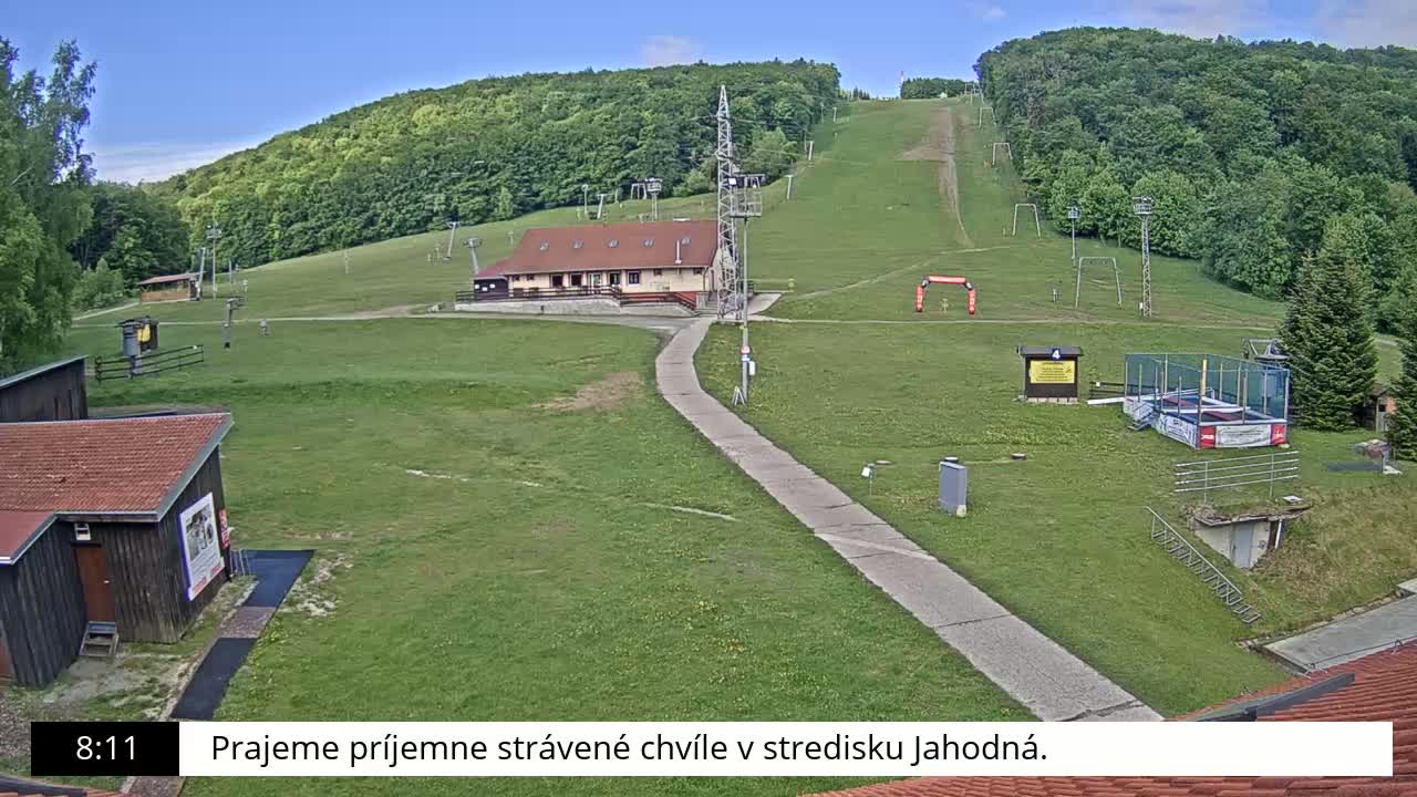 A grassy ski slope with a building, lift infrastructure, and other recreational facilities is visible on a partly sunny day.