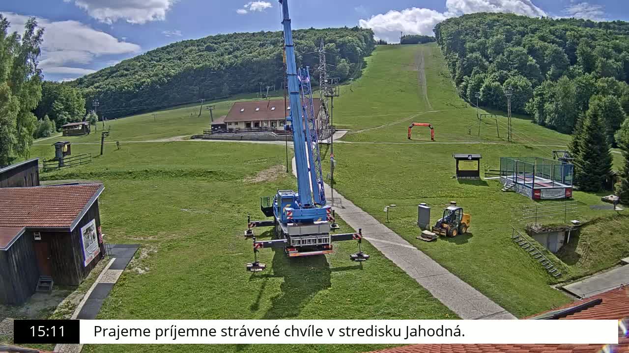 A blue crane is positioned on a grassy area near a small building, with a ski slope and forested hillside visible in the background under a partly cloudy sky.
