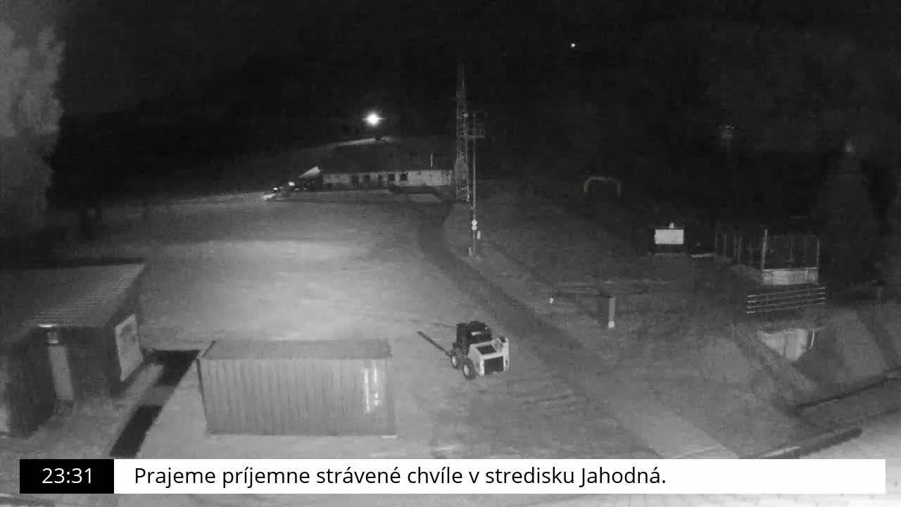 A nighttime, black-and-white view of a mostly empty paved area with a small building, a shipping container, and a piece of machinery.