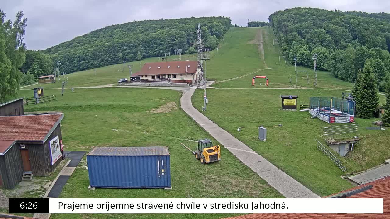 A grassy ski slope with a small building, a bobcat, and a shipping container is visible under an overcast sky.