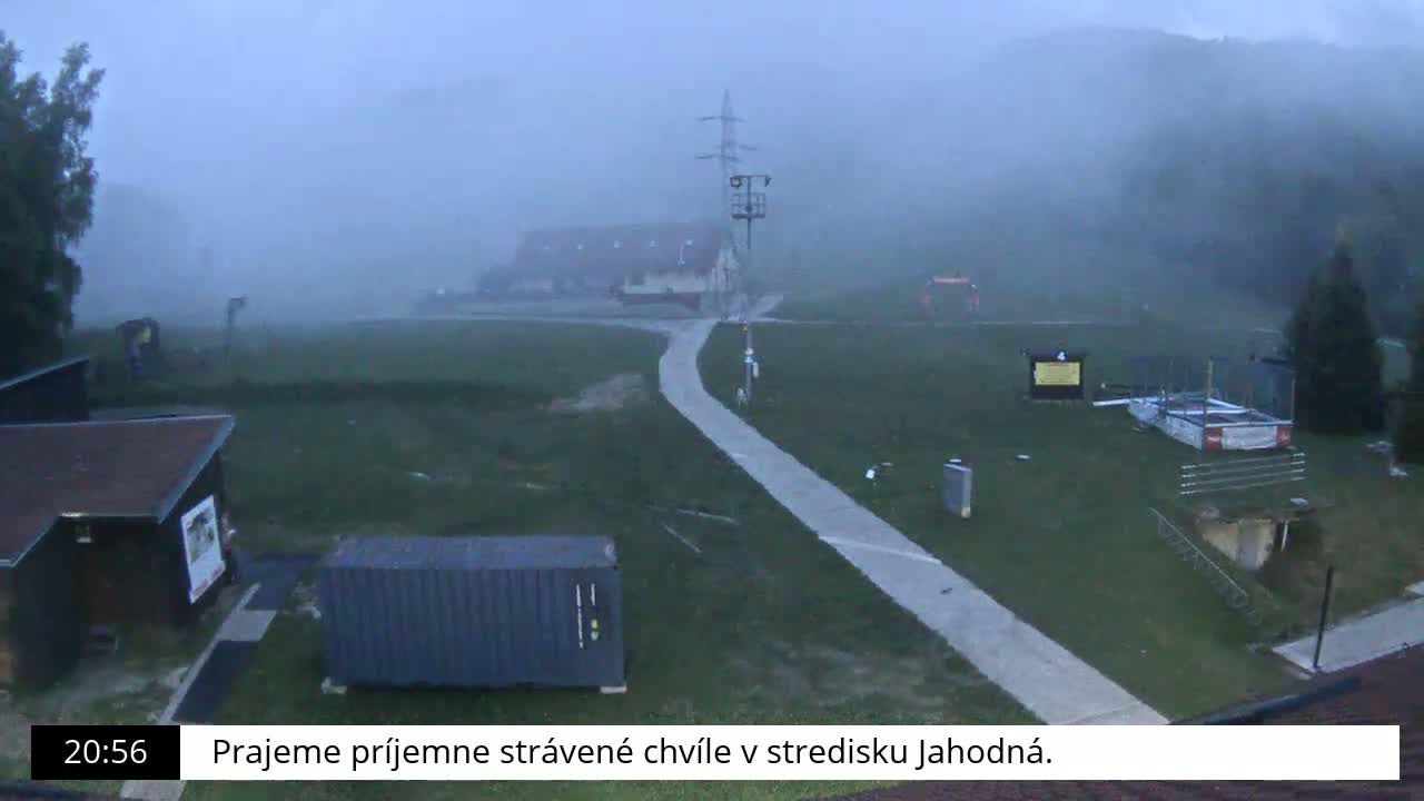 A foggy, outdoor scene shows a paved path leading to a building, surrounded by grassy areas, small structures, and a shipping container.