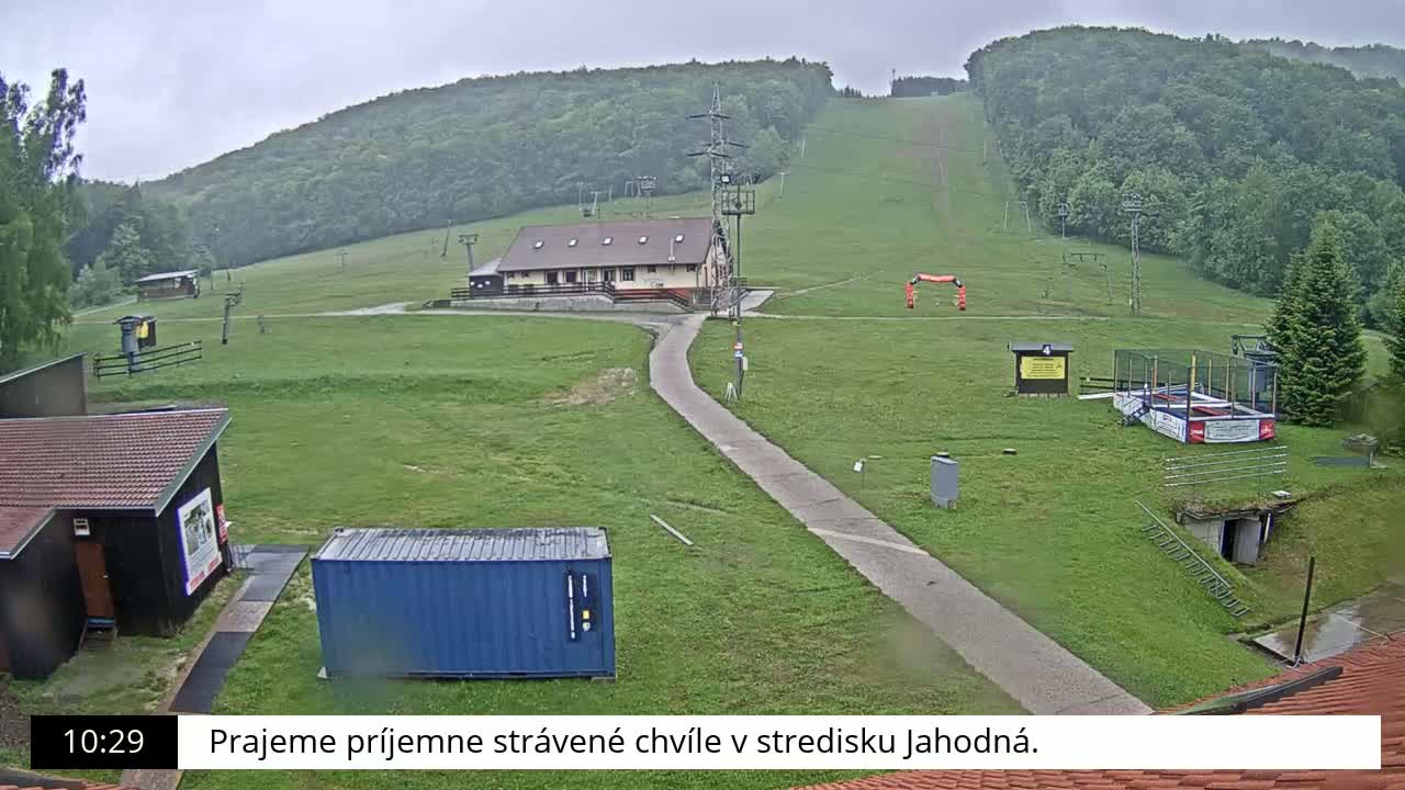 A grassy ski slope with a building, a trampoline, and a blue shipping container is visible on an overcast day.